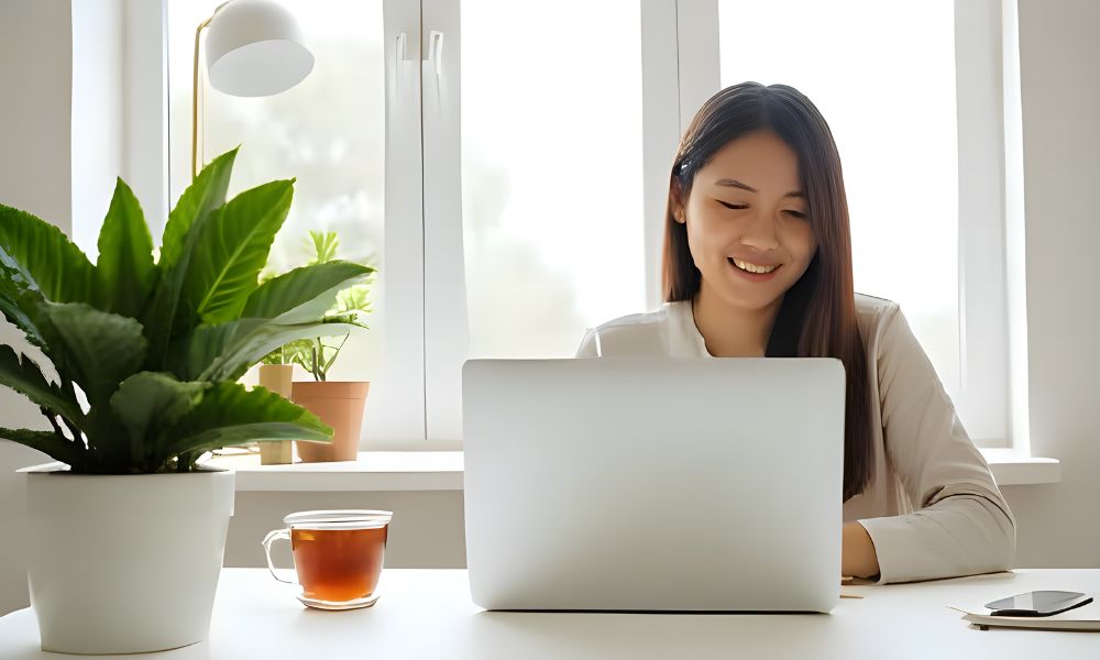 Smiling Asian woman working remotely on a laptop in a bright home office with indoor plants, natural light, and a cup of tea, promoting productive and peaceful remote work environments