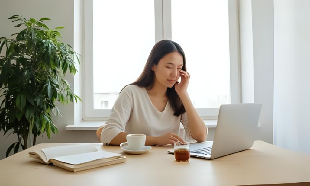 Young Asian woman working on a laptop at home with coffee and books on the table, highlighting remote work flexibility and home office productivity in a bright minimalist setting.