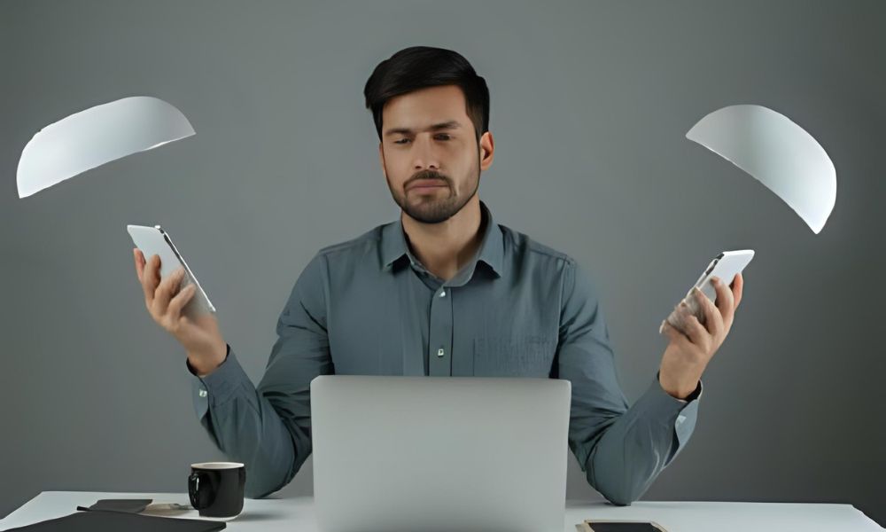 Focused man multitasking with two smartphones and a laptop at a modern desk, symbolizing digital overload and productivity in a tech-driven workspace