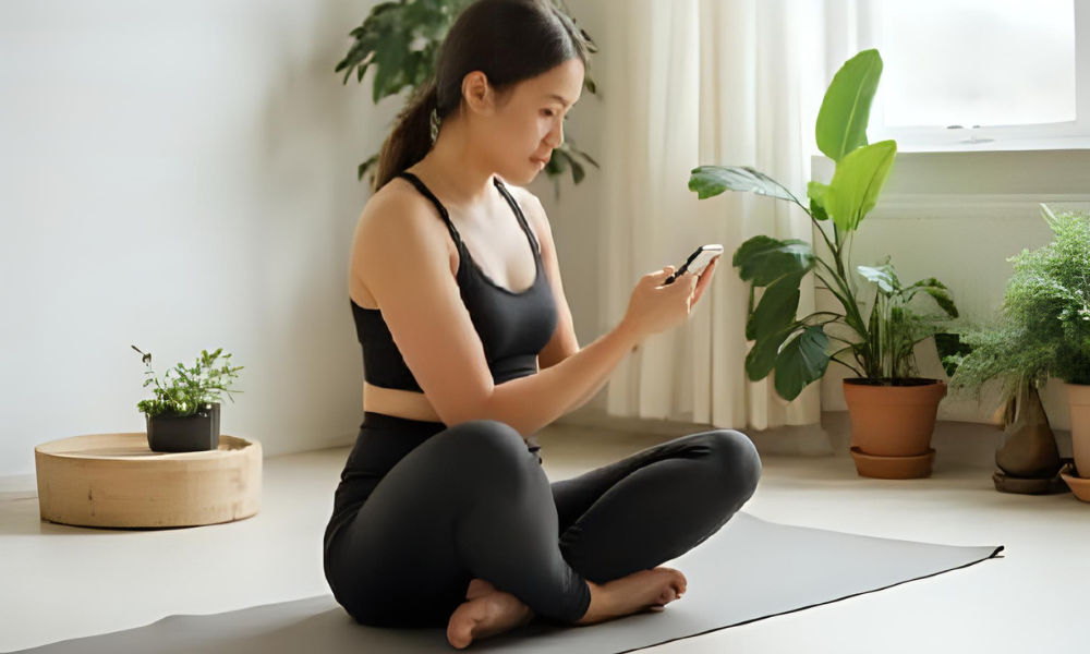 Young Asian woman in activewear sitting cross-legged on a yoga mat, using smartphone in a bright home with indoor plants, representing digital wellness and mindfulness at home