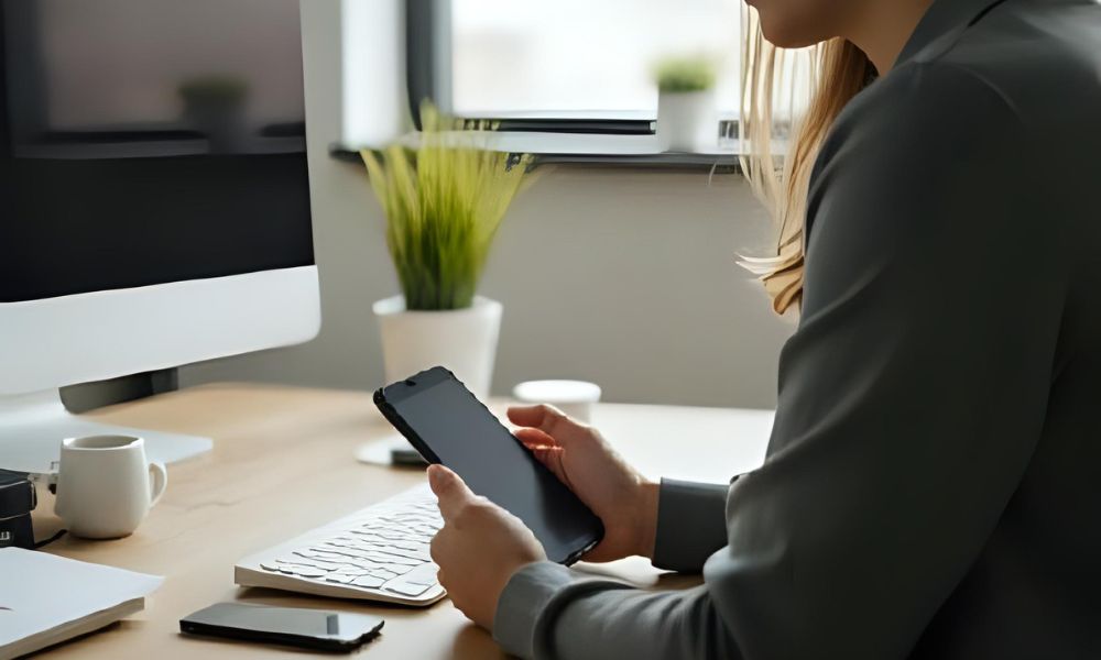 Woman working remotely at a modern desk setup, holding a tablet beside a desktop computer, smartphone, and coffee cup, symbolizing digital productivity and remote work lifestyle