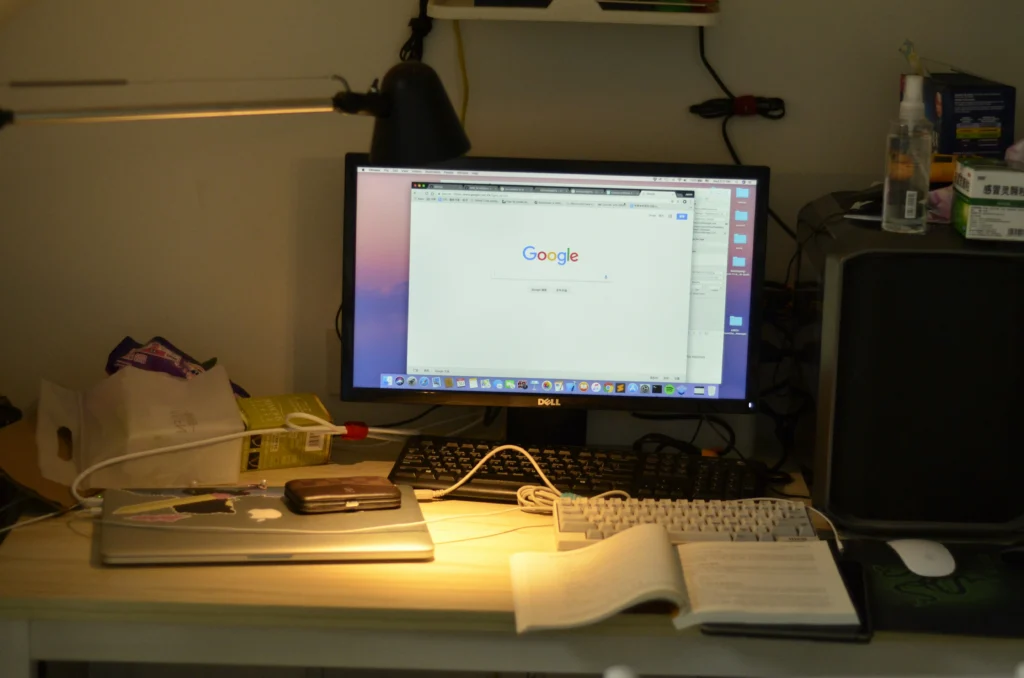 A cluttered desk with a monitor displaying the Google homepage, a mechanical keyboard, laptop, open book, and various items under a lamp. The scene suggests a busy workspace.