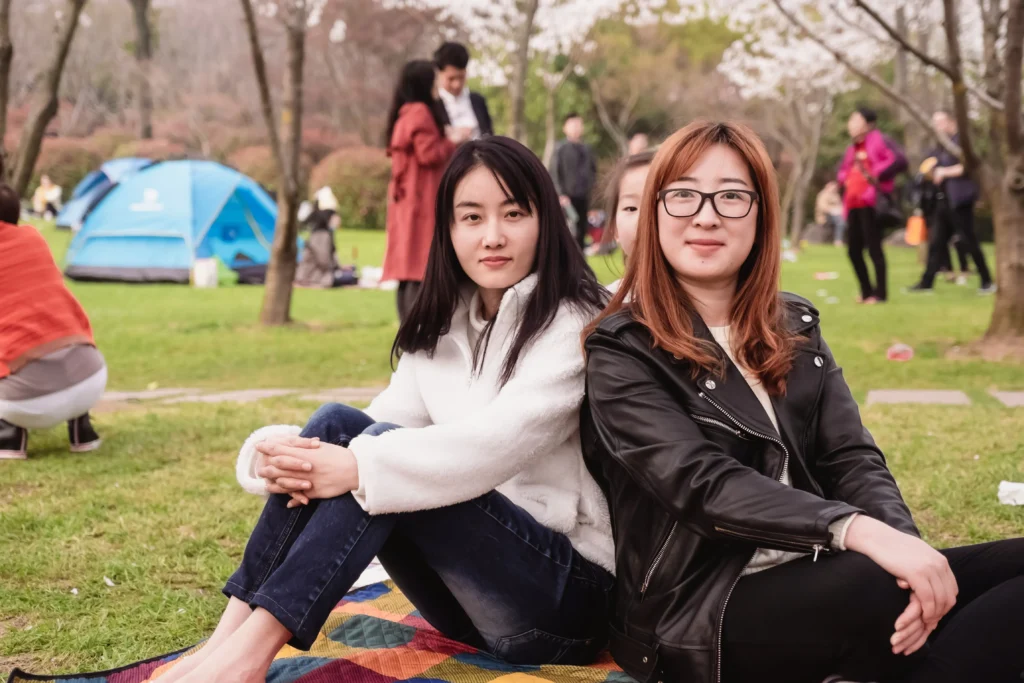 Two women sit back-to-back on a blanket in a park, surrounded by people, trees, and a blue tent in the background.