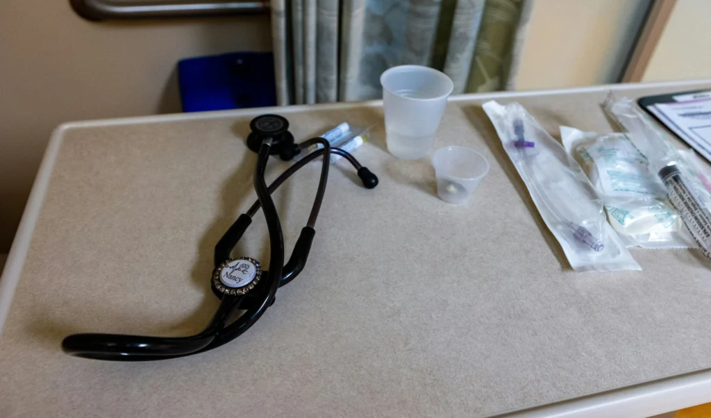 A black stethoscope lies on a beige hospital table alongside medical supplies, including a syringe, alcohol wipes, and small plastic cups, conveying a clinical setting.