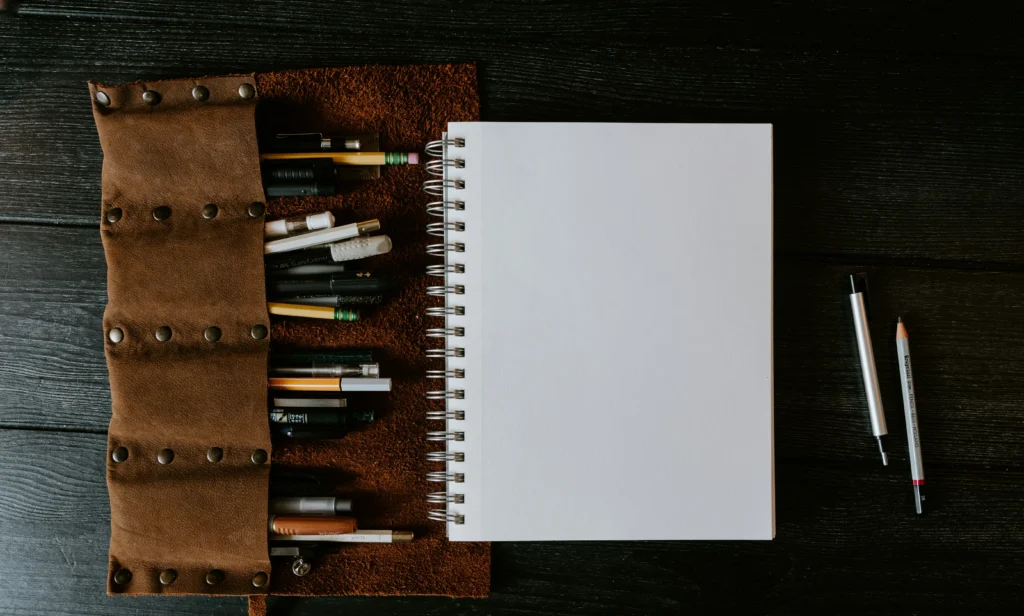 A leather pencil roll with various pens and pencils lies open beside a blank spiral notebook on a dark wooden surface. A pen and pencil are on the right.