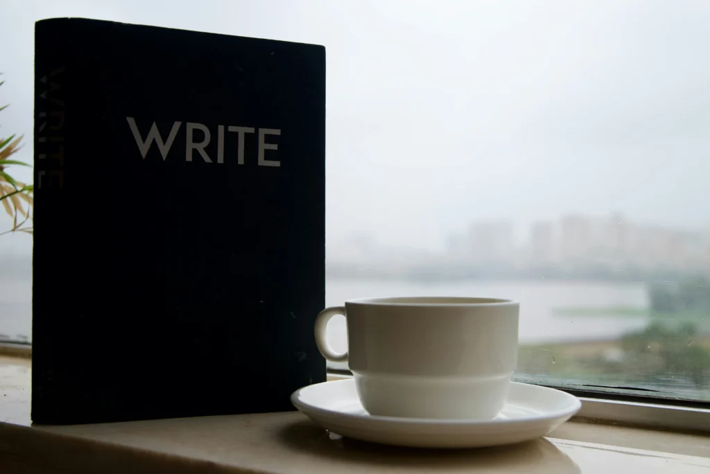 A black book titled "WRITE" sits beside a white cup on a saucer, with a blurred cityscape visible through the window in the background.