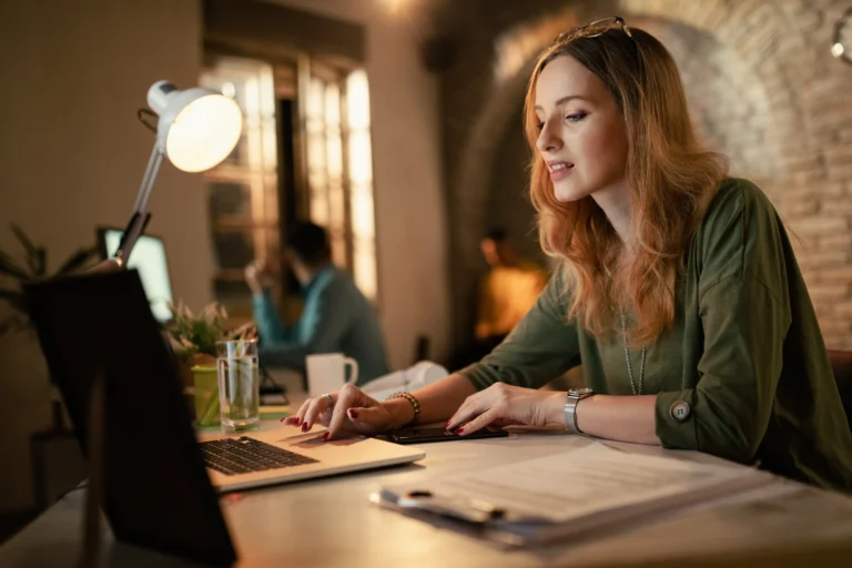 A woman is focused on her laptop while sitting at a desk, surrounded by office supplies and a plant.